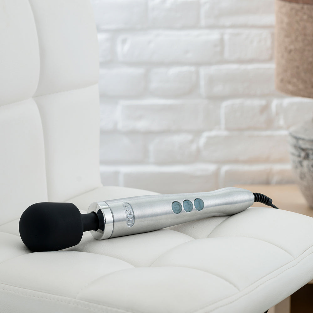 The brushed metal Doxy wand vibrator lying on a couch, blending style with pleasure. Available at SugarX in the Vibrators Collection.