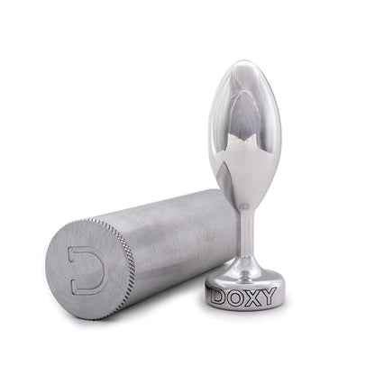 Doxy Smooth Metal Butt Plug standing beside its brushed aluminum storage case which is lying on its side on a plain white background showcasing its tapered aluminum body and extra long stem. Available at SugarX in the Butt Plug section of the Sex Toys col